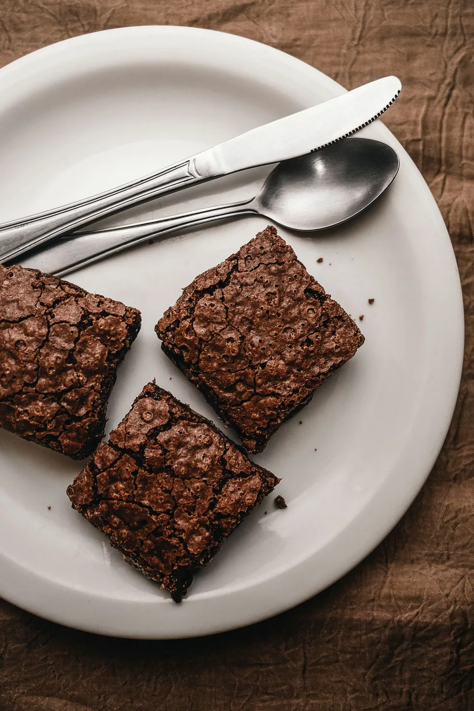Three chocolate brownies on a plate with a knife and spoon