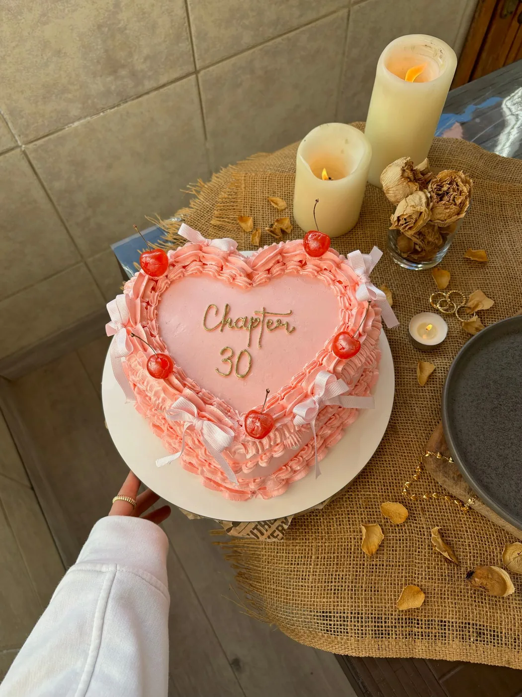 Pink heart-shaped cake with piped ruffles and cherries, held over a table with candles